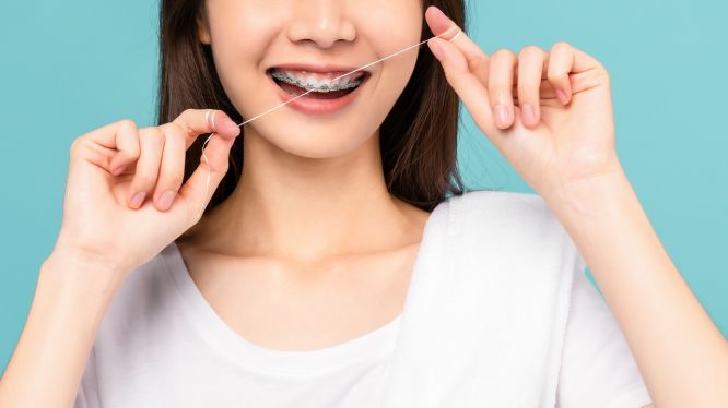 Mujer joven con brackets usando hilo dental frente a un fondo azul, demostrando una técnica adecuada de limpieza interdental durante tratamiento de ortodoncia.